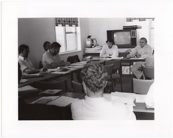 B&W photo of a group of people sitting around some tables placed in a large square formation. There are several documents on every table. In the background, there is a large television set on a stand, with a small table next to it. A briefcase has been placed on top of the television. On top of the table is a large rectangular item with a cover placed over it, an open AIWA (consumer electronics brand) box, and other miscellaneous items. There are also two windows with blinds.