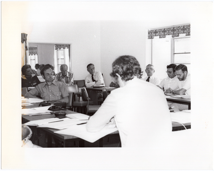 B&W photo of a group of people sitting around some tables placed in a large square formation. One person is sitting on the inside of the square table formation. There are several documents on every table. On one table, there is an ashtray and a square tape recorder with its own cover - one of the operational buttons is depressed.  In the background, there is a large mirror on the wall, on which a reflection of two windows with blinds is evident. Next to the mirror is a piece of fabric hanging over the top of a display board or room divider (partially obscured).