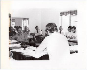 B&W photo of a group of people sitting around some tables placed in a large square formation. One person is sitting on the inside of the square table formation. There are several documents on every table. On one table, there is an ashtray and a square tape recorder with its own cover - one of the operational buttons is depressed.  In the background, there is a large mirror on the wall, on which a reflection of two windows with blinds is evident. Next to the mirror is a piece of fabric hanging over the top of a display board or room divider (partially obscured).