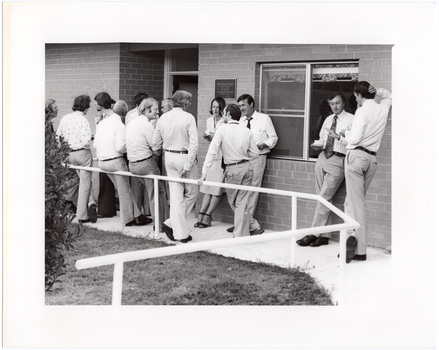 B&W photo of a group of people standing outside a brick building, chatting. Some are holding a cup and saucer in their hands. There is a plaque on the exterior brick wall, containing the text, "RHEUMATISM AND ARTHRITIS ASSOCIATION OF VICTORIA ACTION HOUSE OFFICIALLY OPENED ON ... BY HIS EXCELLENCY THE HONOURABLE SIR HENRY WINNEKE...".