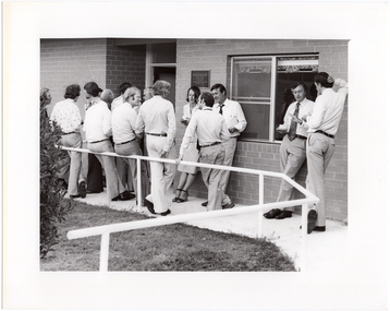 B&W photo of a group of people standing outside a brick building, chatting. Some are holding a cup and saucer in their hands. There is a plaque on the exterior brick wall, containing the text, "RHEUMATISM AND ARTHRITIS ASSOCIATION OF VICTORIA ACTION HOUSE OFFICIALLY OPENED ON ... BY HIS EXCELLENCY THE HONOURABLE SIR HENRY WINNEKE...".