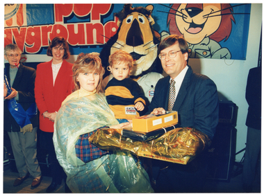 COL photo of a man handing a woman a stack of prizes, some of which have been gift-wrapped. She is holding a toddler in one arm while she accepts the prizes with the other hand. Two people are standing behind, to one side. One of them is wearing a Grandparent of the Year sash across her torso. On the wall behind them, there is a large Streets Ice Cream banner featuring an image of the Paddle Pop Lion and the (partially obscured) text, "POP ...ayground".