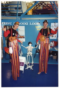 COL photo of two stiltwalers wearing colourful costumes hold hands with a skeleton on a stand. They are at a stall at the Royal Melbourne Show. The wall behind them contains the text "Have a Good Look" and "Your Life... In Your..." (partially obscured). There are some posters on the wall, one of which is titled "MINISTERIAL ADVISORY COUNCIL OF SENIOR VICTORIANS". There is also a small table with various items on it, including a book, some stickers and a water bottle. Behind one of the stiltwalkers is an A-frame sandwich board (partially obscured), with the text, "OSTEOPOROSIS VICTORIA Are you at risk of osteoporosis? ...HERE!". Two other people (partially obscured) are standing in the background.COL photo of two stiltwalers wearing colourful costumes hold hands with a skeleton on a stand. They are at a stall at the Royal Melbourne Show. The wall behind them contains the text "Have a Good Look" and "Your Life... In Your..." (partially obscured). There are some posters on the wall, one of which is titled "MINISTERIAL ADVISORY COUNCIL OF SENIOR VICTORIANS". There is also a small table with various items on it, including a book, some stickers and a water bottle. Behind one of the stiltwalkers is an A-frame sandwich board (partially obscured), with the text "...OPOROSIS TORIA... t risk ...rosis? ...ERE!" Two other people (partially obscured) are standing in the background.