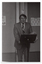 B&W photo of a man standing at a lectern. He has both hands on the lectern, with some papers also in one hand. There are some posters on the wall behind him.