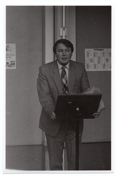 B&W photo of a man standing at a lectern. He has both hands on the lectern, with some papers also in one hand. There are some posters on the wall behind him.