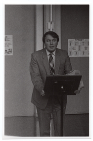 B&W photo of a man standing at a lectern. He has both hands on the lectern, with some papers also in one hand. There are some posters on the wall behind him.