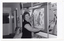 B&W photo of a woman pinning or unpinning an anatomical poster titled, "Common Sites of Arthritis", to/from the centre panel of a display board. On the centre top of the board, the title "RHEUMATISM AND..." (partially obscured) is displayed. On the left side panel, another poster (partially obscured) is titled, "ONE IN FIVE PEOPLE WILL SUFFER FROM ARTHRITIS". The right side panel of the display board (partially obscured) has a range of arthritis aids affixed to it under the title (partially obscured), "AIDS TO...". The centre bottom panel of the board (partially obscurred) is titled, "SURGERY".
