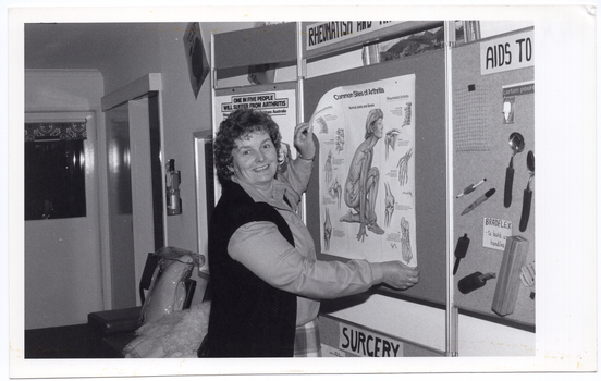 B&W photo of a woman pinning or unpinning an anatomical poster titled, "Common Sites of Arthritis", to/from the centre panel of a display board. On the centre top of the board, the title "RHEUMATISM AND..." (partially obscured) is displayed. On the left side panel, another poster (partially obscured) is titled, "ONE IN FIVE PEOPLE WILL SUFFER FROM ARTHRITIS". The right side panel of the display board (partially obscured) has a range of arthritis aids affixed to it under the title (partially obscured), "AIDS TO...". The centre bottom panel of the board (partially obscurred) is titled, "SURGERY".