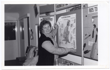 B&W photo of a woman pinning or unpinning an anatomical poster titled, "Common Sites of Arthritis", to/from the centre panel of a display board. On the centre top of the board, the title "RHEUMATISM AND..." (partially obscured) is displayed. On the left side panel, another poster (partially obscured) is titled, "ONE IN FIVE PEOPLE WILL SUFFER FROM ARTHRITIS". The right side panel of the display board (partially obscured) has a range of arthritis aids affixed to it under the title (partially obscured), "AIDS TO...". The centre bottom panel of the board (partially obscurred) is titled, "SURGERY".