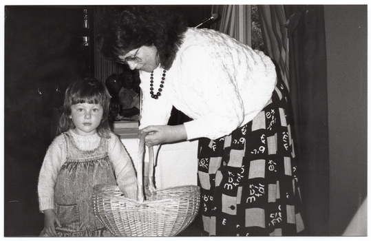 B&W photo of a woman and a young girl. The woman is wearing a light coloured jumber and a dark coloured printed skirt. The girl is wearing a light coloured jumper under a demin pinafore dress. The woman is holding a cane basket and leaning down towards the girl, who has one hand inside the basket. Behind the woman, there is a window with a striped curtain, through which some trees are evident.