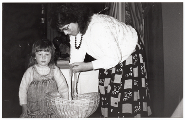 B&W photo of a woman and a young girl. The woman is wearing a light coloured jumber and a dark coloured printed skirt. The girl is wearing a light coloured jumper under a demin pinafore dress. The woman is holding a cane basket and leaning down towards the girl, who has one hand inside the basket. Behind the woman, there is a window with a striped curtain, through which some trees are evident.