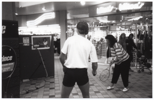 B&W photo of two people, each holding a tennis racket, practising serving tennis balls into a small framed net. Behind the net is a display board with a poster of a woman playing tennis. To the left of the frame, there is another display board advertising Prince tennis products. In the background, there are two people walking past shops with neon signage. One of them is pushing a pram. They are both looking towards the tennis players.