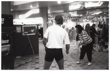 B&W photo of two people, each holding a tennis racket, practising serving tennis balls into a small framed net. Behind the net is a display board with a poster of a woman playing tennis. To the left of the frame, there is another display board advertising Prince tennis products. In the background, there are two people walking past shops with neon signage. One of them is pushing a pram. They are both looking towards the tennis players.