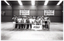 B&W photo of a large group of people posing for a photo behind the net on an indoor tennis court.
