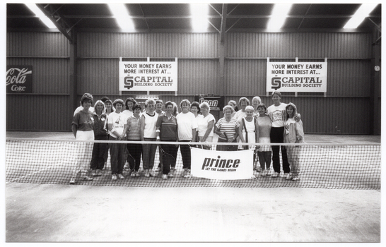B&W photo of a large group of people posing for a photo behind the net on an indoor tennis court.