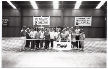 B&W photo of a large group of people posing for a photo behind the net on an indoor tennis court.