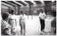 B&W photo of five women standing on one of two indoor tennis courts. They are each holding a tennis racket. One woman is demonstrating how to hold the racket while the other four women are watching her. In the background, there are some advertising signs on the wall, including two for Prince tennis products, one for Coca-Cola soft drinks, and one for a building society (financial institution). The text on that sign (partially obscured) says, "YOUR MONEY MORE INTEREST CAPITOL BUILDING SOCIETY".