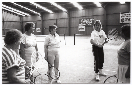 B&W photo of five women standing on one of two indoor tennis courts. They are each holding a tennis racket. One woman is demonstrating how to hold the racket while the other four women are watching her. In the background, there are some advertising signs on the wall, including two for Prince tennis products, one for Coca-Cola soft drinks, and one for a building society (financial institution). The text on that sign (partially obscured) says, "YOUR MONEY MORE INTEREST CAPITOL BUILDING SOCIETY".