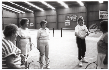 B&W photo of five women standing on one of two indoor tennis courts. They are each holding a tennis racket. One woman is demonstrating how to hold the racket while the other four women are watching her. In the background, there are some advertising signs on the wall, including two for Prince tennis products, one for Coca-Cola soft drinks, and one for a building society (financial institution). The text on that sign (partially obscured) says, "YOUR MONEY MORE INTEREST CAPITOL BUILDING SOCIETY".