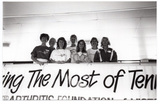B&W photo of a small group of women standing behind a barrier on a viewing platform or balcony. On the barricade, there is a large banner (partially obscured) with the text, "Making The Most of Tenhis" and "... ARTHRITIS FOUNDATION of VICTORIA" (partially obscured).