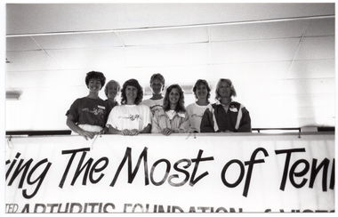 B&W photo of a small group of women standing behind a barrier on a viewing platform or balcony. On the barricade, there is a large banner (partially obscured) with the text, "Making The Most of Tenhis" and "... ARTHRITIS FOUNDATION of VICTORIA" (partially obscured).