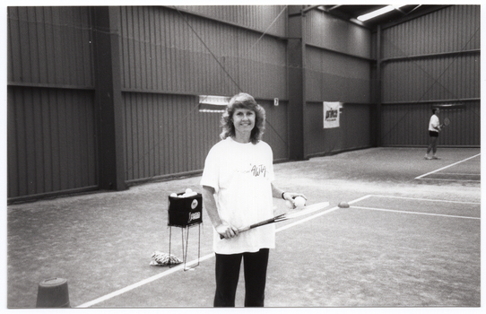 B&W photo of a woman standing on an indoor tennis court. She is wearing a t-shirt with the logo for the Australian Women's Tennis Association. She is holding two tennis balls in one hand, poised over a tennis racket which she is holding with her other hand. Behind her, there is a square container on a stand, filled with tennis balls. In the background, to the right of the frame, a man is playing tennis on an adjoining court. On the wall, there is an advertising sign for Prince tennis products.