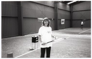 B&W photo of a woman standing on an indoor tennis court. She is wearing a t-shirt with the logo for the Australian Women's Tennis Association. She is holding two tennis balls in one hand, poised over a tennis racket which she is holding with her other hand. Behind her, there is a square container on a stand, filled with tennis balls. In the background, to the right of the frame, a man is playing tennis on an adjoining court. On the wall, there is an advertising sign for Prince tennis products.