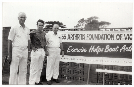 B&W photo of three men standing next to a large horizontal banner affixed to an exterior wall constructed from decorative concrete blocks (also known as "breeze blocks"). At the top of the banner, in large font, is the text (partially obscurred), "ARTHRITIS FOUNDATION OF VICTORIA", preceeded by a telephone number. Across the centre, there is the text, "Exercise Helps Beat Arthritis" (partially obscured). Across the bottom of the banner, there is the text (partially obscured), "...ER PROJECT FUNDED BY VICTORIAN HEALTH PROMOTION FOUNDATION”.