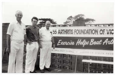 B&W photo of three men standing next to a large horizontal banner affixed to an exterior wall constructed from decorative concrete blocks (also known as "breeze blocks"). At the top of the banner, in large font, is the text (partially obscurred), "ARTHRITIS FOUNDATION OF VICTORIA", preceeded by a telephone number. Across the centre, there is the text, "Exercise Helps Beat Arthritis" (partially obscured). Across the bottom of the banner, there is the text (partially obscured), "...ER PROJECT FUNDED BY VICTORIAN HEALTH PROMOTION FOUNDATION”.
