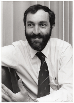 B&W close up photo of Dr Tim Woodruff. He is sitting at a slight angle on a row of chairs. Behind him, there are some vertical venetian blinds covering a window.