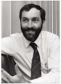 B&W close up photo of Dr Tim Woodruff. He is sitting at a slight angle on a row of chairs. Behind him, there are some vertical venetian blinds covering a window.