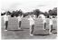 B&W photo of a group of women, standing slightly apart, on a grassy oval. They are wearing sweatshirts with the "Joint Action Group" logo. They each have their arms outstretched to the side. In the distance, there are several tents with people out the front, and some vehicles parked nearby.  Behind the tents, people, and cars, there is a row of large trees.
