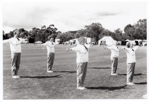 B&W photo of a group of women, standing slightly apart, on a grassy oval. They are wearing sweatshirts with the "Joint Action Group" logo. They each have their arms outstretched to the side. In the distance, there are several tents with people out the front, and some vehicles parked nearby.  Behind the tents, people, and cars, there is a row of large trees.