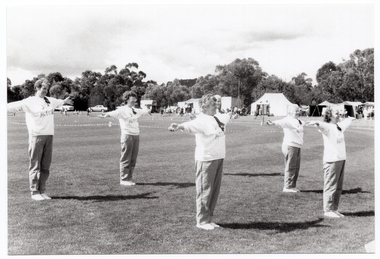 B&W photo of a group of women, standing slightly apart, on a grassy oval. They are wearing sweatshirts with the "Joint Action Group" logo. They each have their arms outstretched to the side. In the distance, there are several tents with people out the front, and some vehicles parked nearby.  Behind the tents, people, and cars, there is a row of large trees.