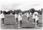 B&W photo of a group of women, standing slightly apart, on a grassy oval. They are wearing sweatshirts with the "Joint Action Group" logo. Most are standing with their arms by their sides. One woman has one hand on her shoulder.  In the distance, there are several tents with people out the front, and some vehicles parked nearby.  Behind the tents, people, and cars, there is a row of large trees.