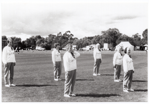 B&W photo of a group of women, standing slightly apart, on a grassy oval. They are wearing sweatshirts with the "Joint Action Group" logo. Most are standing with their arms by their sides. One woman has one hand on her shoulder.  In the distance, there are several tents with people out the front, and some vehicles parked nearby.  Behind the tents, people, and cars, there is a row of large trees.