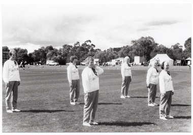 B&W photo of a group of women, standing slightly apart, on a grassy oval. They are wearing sweatshirts with the "Joint Action Group" logo. Most are standing with their arms by their sides. One woman has one hand on her shoulder.  In the distance, there are several tents with people out the front, and some vehicles parked nearby.  Behind the tents, people, and cars, there is a row of large trees.