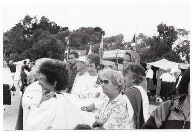 B&W photo of a small group of people standing together while looking out beyond the frame of the photo. Some of them are wearing 'Joint Action Group/Move it or Lose it' windcheaters/sweatshirts. One woman is wearing a tartan scarf over her shoulder and, in the background, people wearing kilts in the same or similar tartan check can be seen. There are some tents in the background. One of these is displaying a banner with the text (partially obscured), "MORNINGTON PENINSULA CALEDONIAN SOCIETY". There are several large trees behind the row of market tents.