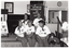 B&W photo of a small group of women posing for a photo while sitting on the edge of a small stage. Each woman is wearing a t-shirt with a "Joint Action" logo. Another woman (partially obscured), also wearing the same "Joint Action" branded t-shirt, is standing on the right side of the frame in front of a small table. On the stage, there is an A-frame sandwich board (partially obscured) with the text and logo for the Arthritis Foundation of Victoria, followed by the same "Joint Action" graphic text illustrated as bones and/or body parts making up the letters. Also on the stage is a vertical 'Arthritis Foundation of Victoria' banner. On the wall, at the rear of the stage, is a portrait of Queen Elizabeth II (1926-2022), a framed print (partially obscured), and a clock.