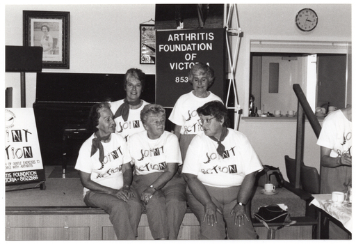 B&W photo of a small group of women posing for a photo while sitting on the edge of a small stage. Each woman is wearing a t-shirt with a "Joint Action" logo. Another woman (partially obscured), also wearing the same "Joint Action" branded t-shirt, is standing on the right side of the frame in front of a small table. On the stage, there is an A-frame sandwich board (partially obscured) with the text and logo for the Arthritis Foundation of Victoria, followed by the same "Joint Action" graphic text illustrated as bones and/or body parts making up the letters. Also on the stage is a vertical 'Arthritis Foundation of Victoria' banner. On the wall, at the rear of the stage, is a portrait of Queen Elizabeth II (1926-2022), a framed print (partially obscured), and a clock.