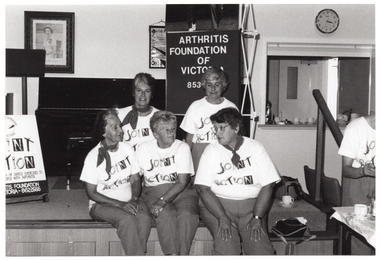 B&W photo of a small group of women posing for a photo while sitting on the edge of a small stage. Each woman is wearing a t-shirt with a "Joint Action" logo. Another woman (partially obscured), also wearing the same "Joint Action" branded t-shirt, is standing on the right side of the frame in front of a small table. On the stage, there is an A-frame sandwich board (partially obscured) with the text and logo for the Arthritis Foundation of Victoria, followed by the same "Joint Action" graphic text illustrated as bones and/or body parts making up the letters. Also on the stage is a vertical 'Arthritis Foundation of Victoria' banner. On the wall, at the rear of the stage, is a portrait of Queen Elizabeth II (1926-2022), a framed print (partially obscured), and a clock.