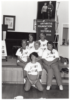 B&W photo of a small group of women posing for a photo in front of a small stage. Five of the women are sitting on the edge of the stage, while one woman is leaning back on her haunches in front of them. Each woman is wearing a t-shirt with a "Joint Action" logo. On the stage, there is an A-frame sandwich board (partially obscured) with the text and logo for the Arthritis Foundation of Victoria, and the letters for "Joint Action" illustrated as bones and/or body parts. Also on the stage is a vertical Arthritis Foundation of Victoria banner and, above it, a large photo poster of two people walking. On the wall at the rear of the stage is a portrait of Queen Elizabeth II (1926-2022) and a framed print.