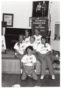 B&W photo of a small group of women posing for a photo in front of a small stage. Five of the women are sitting on the edge of the stage, while one woman is leaning back on her haunches in front of them. Each woman is wearing a t-shirt with a "Joint Action" logo. On the stage, there is an A-frame sandwich board (partially obscured) with the text and logo for the Arthritis Foundation of Victoria, and the letters for "Joint Action" illustrated as bones and/or body parts. Also on the stage is a vertical Arthritis Foundation of Victoria banner and, above it, a large photo poster of two people walking. On the wall at the rear of the stage is a portrait of Queen Elizabeth II (1926-2022) and a framed print.