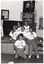 B&W photo of a small group of women posing for a photo in front of a small stage. Five of the women are sitting on the edge of the stage, while one woman is leaning back on her haunches in front of them. Each woman is wearing a t-shirt with a "Joint Action" logo. On the stage, there is an A-frame sandwich board (partially obscured) with the text and logo for the Arthritis Foundation of Victoria, and the letters for "Joint Action" illustrated as bones and/or body parts. Also on the stage is a vertical 'Arthritis Foundation of Victoria' banner with a large photo of two people walking on the top panel. On the wall at the rear of the stage, there is a portrait of Queen Elizabeth II (1926-2022) and a framed print.