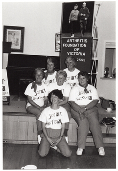 B&W photo of a small group of women posing for a photo in front of a small stage. Five of the women are sitting on the edge of the stage, while one woman is leaning back on her haunches in front of them. Each woman is wearing a t-shirt with a "Joint Action" logo. On the stage, there is an A-frame sandwich board (partially obscured) with the text and logo for the Arthritis Foundation of Victoria, and the letters for "Joint Action" illustrated as bones and/or body parts. Also on the stage is a vertical 'Arthritis Foundation of Victoria' banner with a large photo of two people walking on the top panel. On the wall at the rear of the stage, there is a portrait of Queen Elizabeth II (1926-2022) and a framed print.