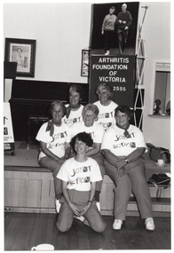 B&W photo of a small group of women posing for a photo in front of a small stage. Five of the women are sitting on the edge of the stage, while one woman is leaning back on her haunches in front of them. Each woman is wearing a t-shirt with a "Joint Action" logo. On the stage, there is an A-frame sandwich board (partially obscured) with the text and logo for the Arthritis Foundation of Victoria, and the letters for "Joint Action" illustrated as bones and/or body parts. Also on the stage is a vertical 'Arthritis Foundation of Victoria' banner with a large photo of two people walking on the top panel. On the wall at the rear of the stage, there is a portrait of Queen Elizabeth II (1926-2022) and a framed print.