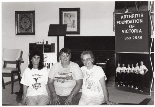 B&W photo of three women posing for a photo, sitting on the edge of a stage. One woman is wearing a VicHealth Masters Games t-shirt; another woman is wearing a Yarrawonga Lake Mulwala 1989 t-shirt. On the stage, there is a vertical Arthritis Foundation of Victoria banner with a large photo poster of the Joint Action Group on the lower panel. On the wall at the rear of the stage, there is a portrait of Queen Elizabeth II (1926-2022) and two framed tapestries. On the stage, there is a piano, some electronic equipment with an antenna on top of a small cabinet, a chair, and an A-frame sandwich board with the "Joint Action" logo on it.