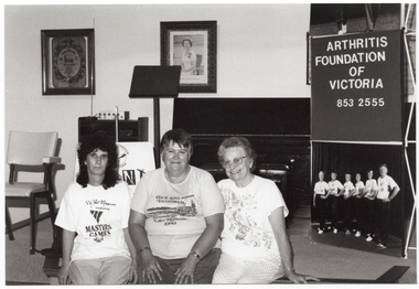 B&W photo of three women posing for a photo, sitting on the edge of a stage. One woman is wearing a VicHealth Masters Games t-shirt; another woman is wearing a Yarrawonga Lake Mulwala 1989 t-shirt. On the stage, there is a vertical Arthritis Foundation of Victoria banner with a large photo poster of the Joint Action Group on the lower panel. On the wall at the rear of the stage, there is a portrait of Queen Elizabeth II (1926-2022) and two framed tapestries. On the stage, there is a piano, some electronic equipment with an antenna on top of a small cabinet, a chair, and an A-frame sandwich board with the "Joint Action" logo on it.