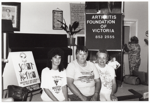 B&W photo of three women posing for a photo, sitting on the edge of a stage. One woman is wearing a VicHealth Masters Games t-shirt; another woman is wearing a Yarrawonga Lake Mulwala 1989 t-shirt. On the stage, there is a vertical Arthritis Foundation of Victoria banner with a large photo poster of the Joint Action Group (partially obscured) on the lower panel. On the wall at the rear of the stage, there is a portrait of Queen Elizabeth II (1926-2022) and two framed tapestries. On the stage, there is a piano, a potted plant on a stand, a lectern, and an A-frame sandwich board with the "Joint Action" logo on it, and a shoulder bag. In the background, another woman is standing in front of an open servery. This photo has been taken from the left of the subjects, at a slight angle.