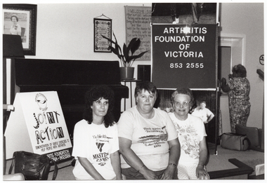 B&W photo of three women posing for a photo, sitting on the edge of a stage. One woman is wearing a VicHealth Masters Games t-shirt; another woman is wearing a Yarrawonga Lake Mulwala 1989 t-shirt. On the stage, there is a vertical Arthritis Foundation of Victoria banner with a large photo poster of the Joint Action Group (partially obscured) on the lower panel. On the wall at the rear of the stage, there is a portrait of Queen Elizabeth II (1926-2022) and two framed tapestries. On the stage, there is a piano, a potted plant on a stand, a lectern, and an A-frame sandwich board with the "Joint Action" logo on it, and a shoulder bag. In the background, another woman is standing in front of an open servery. This photo has been taken from the left of the subjects, at a slight angle.